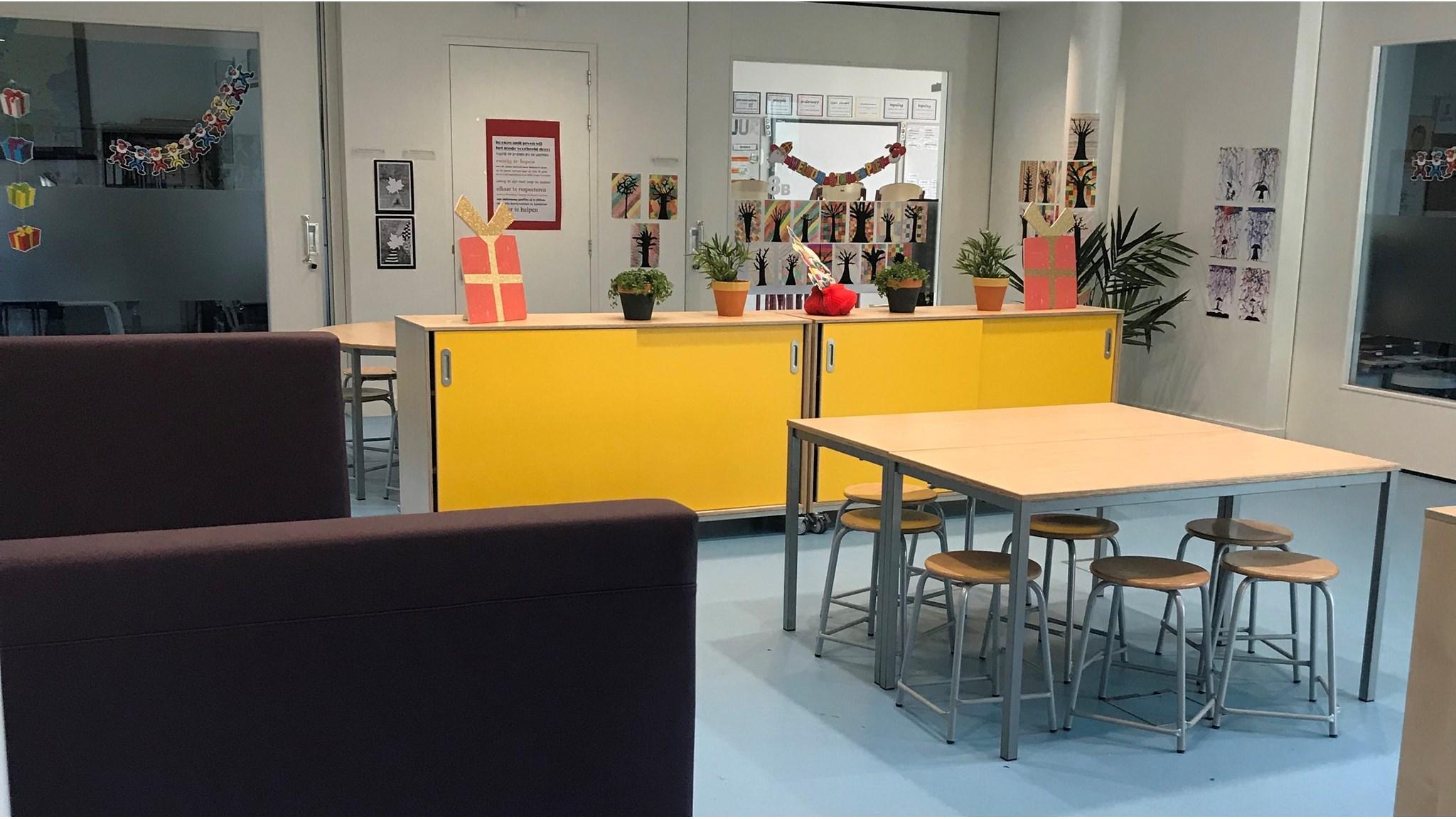 De afbeelding toont een kleurrijke lesruimte van Rk Bs De Mheyster in Hoensbroek met een gele kast en kunstwerken van leerlingen. De afbeelding toont een kleurrijke lesruimte van Rk Bs De Mheyster in Hoensbroek met een gele kast en kunstwerken van leerlingen.
