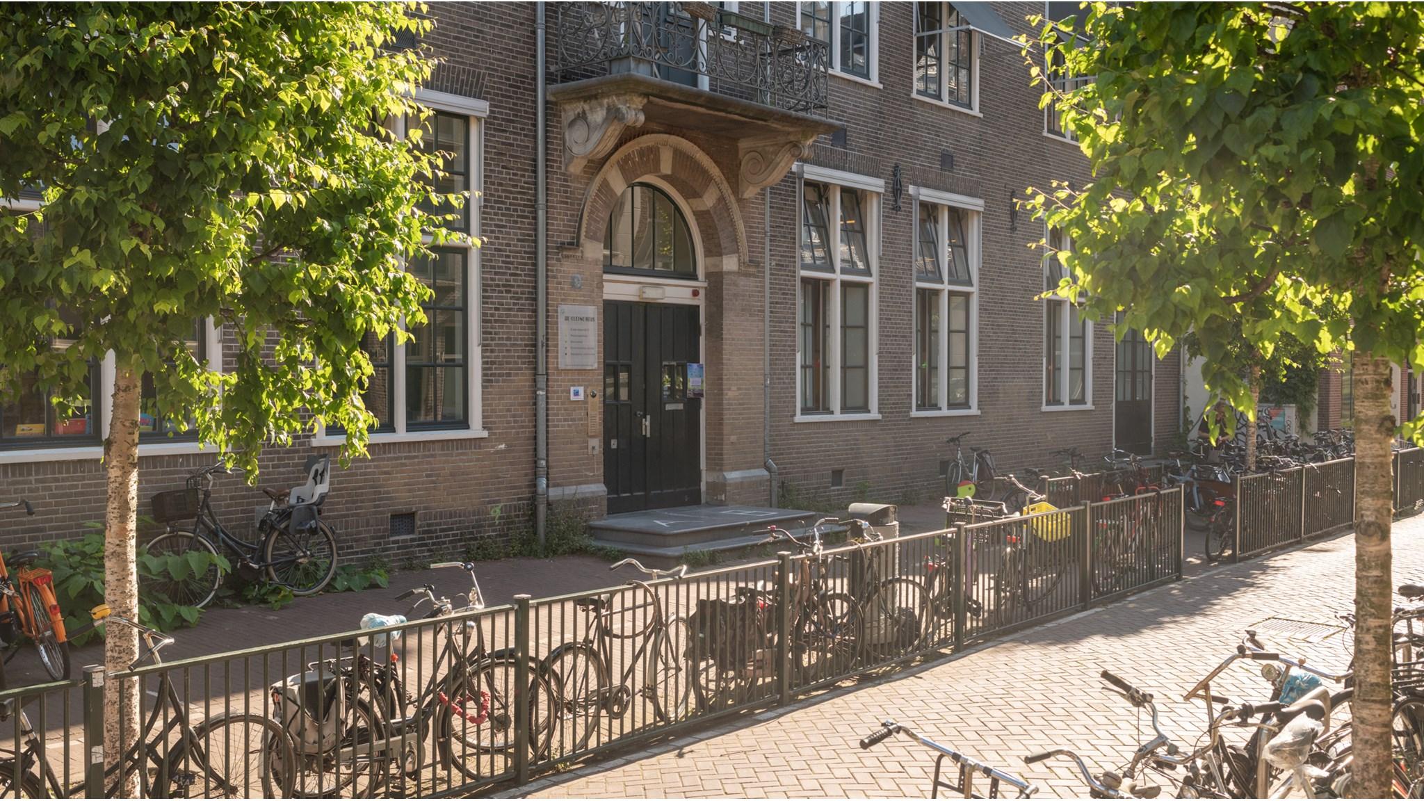 De gevel van IKC De Kleine Reus in Amsterdam met fietsen en groene bomen op de voorgrond. De gevel van IKC De Kleine Reus in Amsterdam met fietsen en groene bomen op de voorgrond.