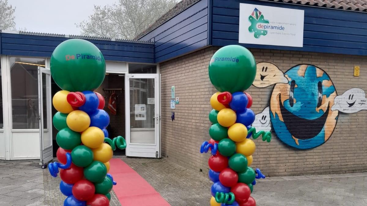 De ingang van Openbare Basisschool De Piramide in Edam, versierd met ballonnen en een kleurrijke muurkunst. De ingang van Openbare Basisschool De Piramide in Edam, versierd met ballonnen en een kleurrijke muurkunst.