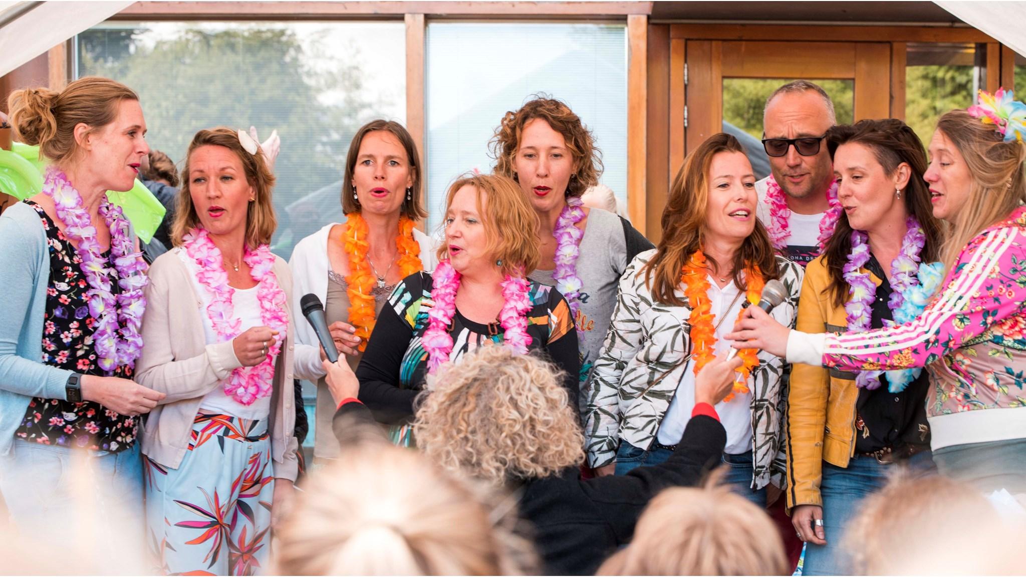 Deze foto toont een viering op OBS Twiske in Amsterdam, waarbij een groep vrouwen met bloemenkransen samen zingt. Deze foto toont een viering op OBS Twiske in Amsterdam, waarbij een groep vrouwen met bloemenkransen samen zingt.