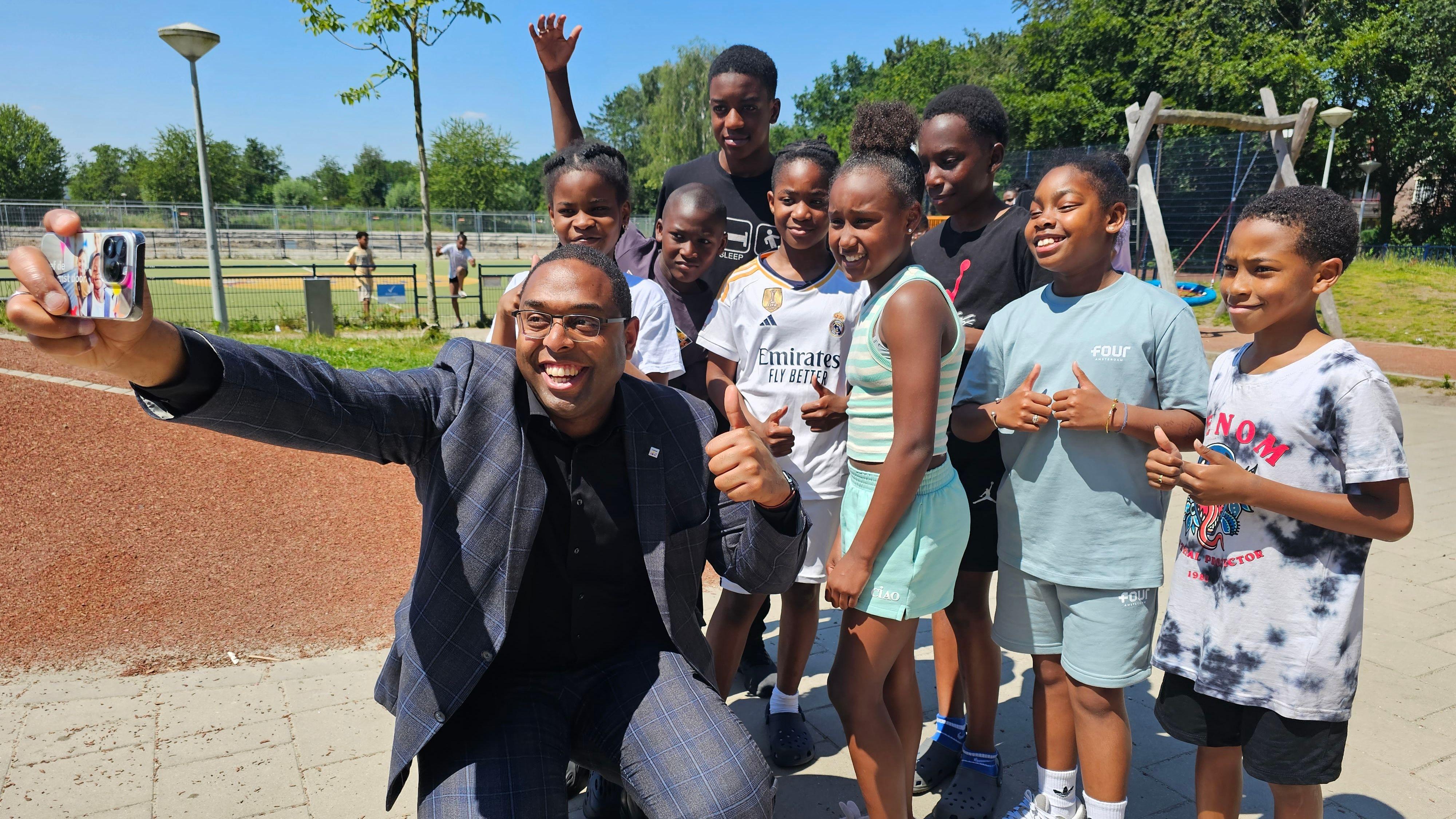 Een groep enthousiaste kinderen poseert met een volwassene voor een selfie op het schoolplein van IKC Knotwilg in Amsterdam. Een groep enthousiaste kinderen poseert met een volwassene voor een selfie op het schoolplein van IKC Knotwilg in Amsterdam.