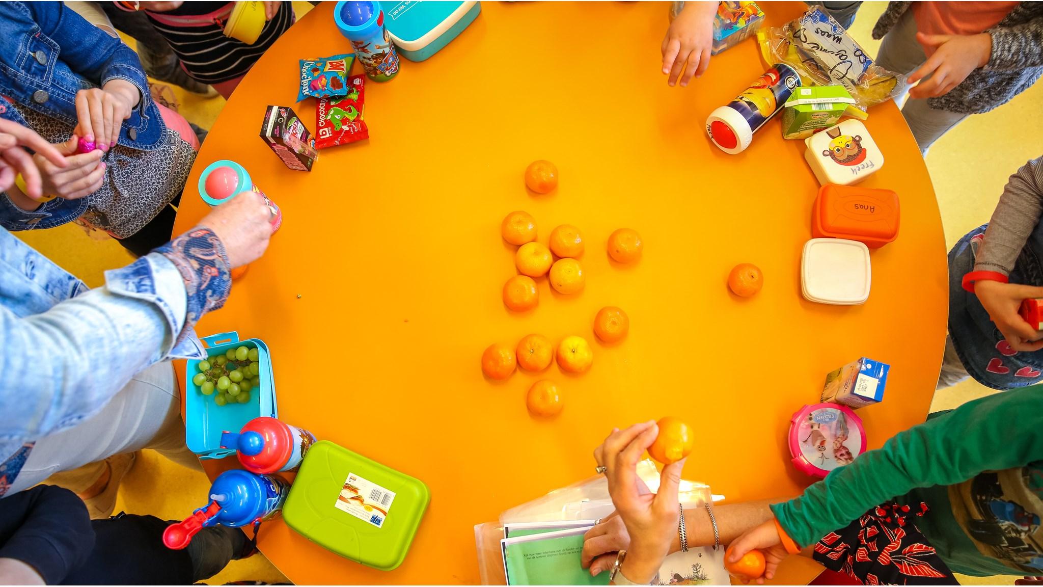 Een groep kinderen van Basisschool De Windroos in Valkenswaard geniet van hun fruitpauze aan een feloranje tafel. Een groep kinderen van Basisschool De Windroos in Valkenswaard geniet van hun fruitpauze aan een feloranje tafel.