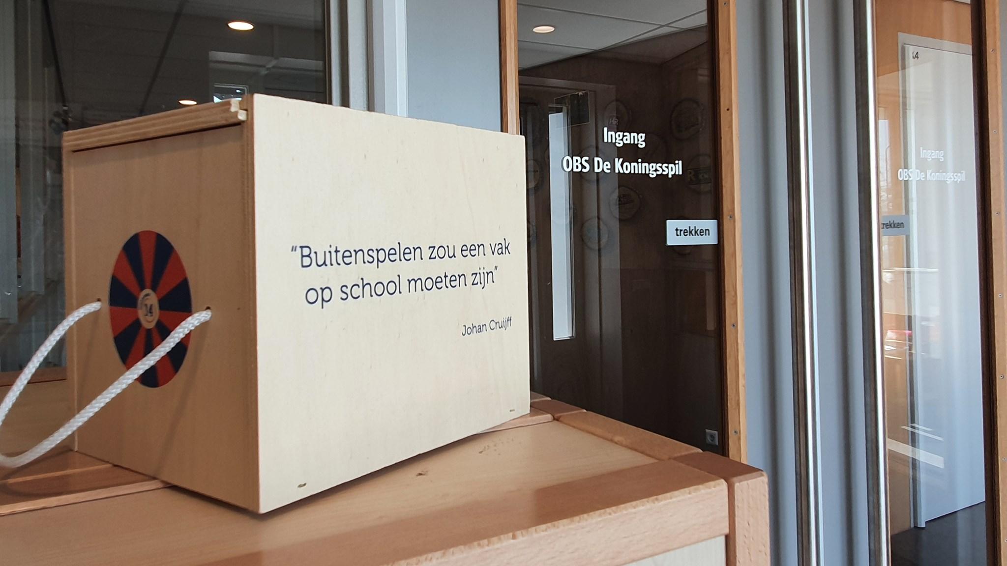 Een houten doos met de tekst "Buitenspelen zou een vak op school moeten zijn" van Johan Cruijff, bij de ingang van Openbare Basisschool De Koningsspil in Oosthuizen.