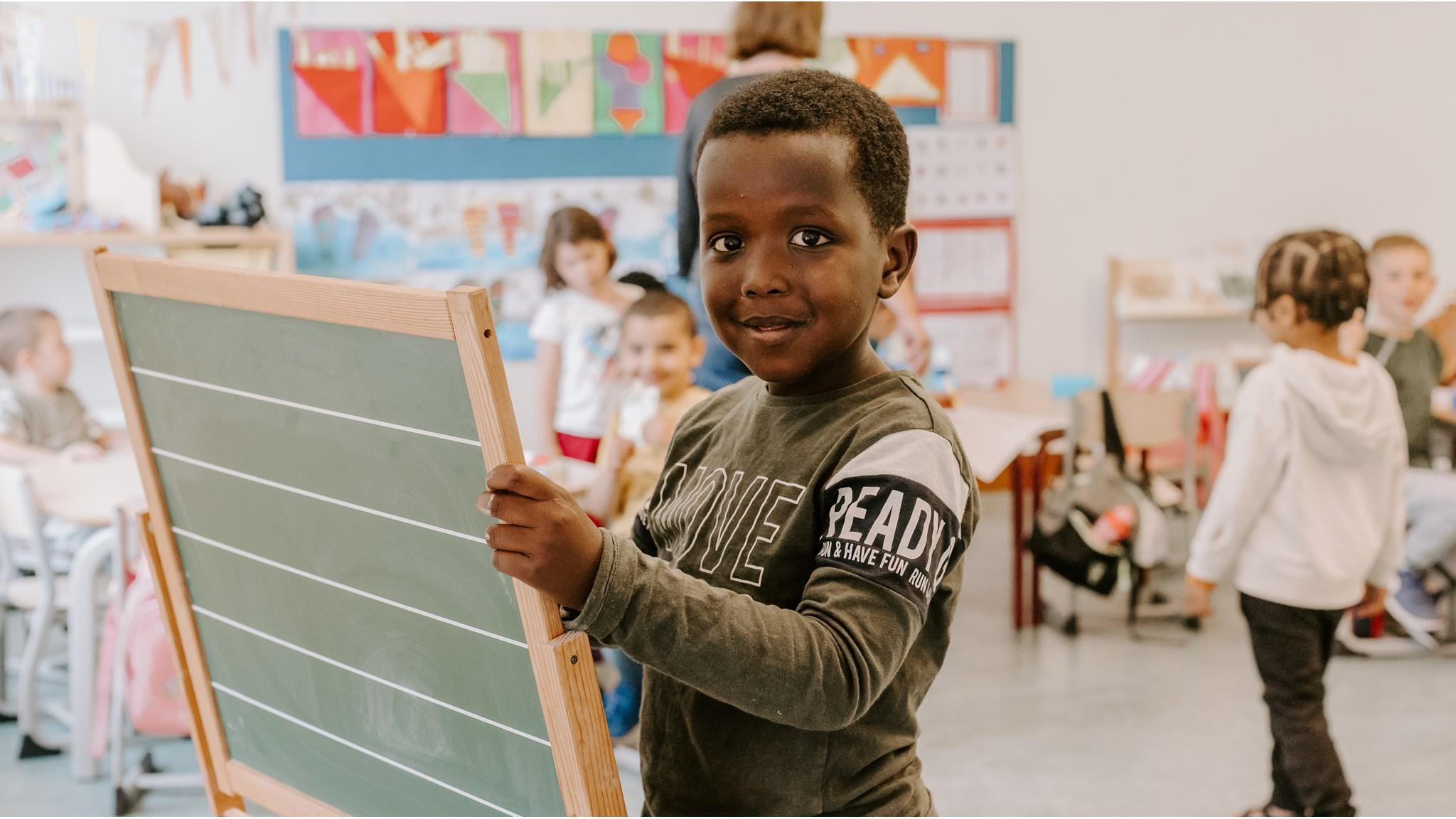 Een jongen in de klas van de Prinses Marijkeschool in Axel houdt een schoolbord vast terwijl andere kinderen op de achtergrond spelen. Een jongen in de klas van de Prinses Marijkeschool in Axel houdt een schoolbord vast terwijl andere kinderen op de achtergrond spelen.