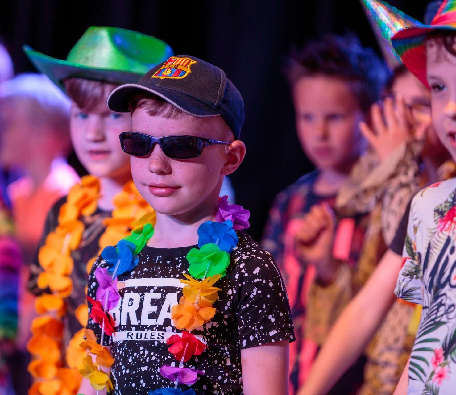Een jongen met een hoed en zonnebril staat op het podium tijdens een feestelijke gebeurtenis op de Antoniusschool in Kudelstaart. Een jongen met een hoed en zonnebril staat op het podium tijdens een feestelijke gebeurtenis op de Antoniusschool in Kudelstaart.