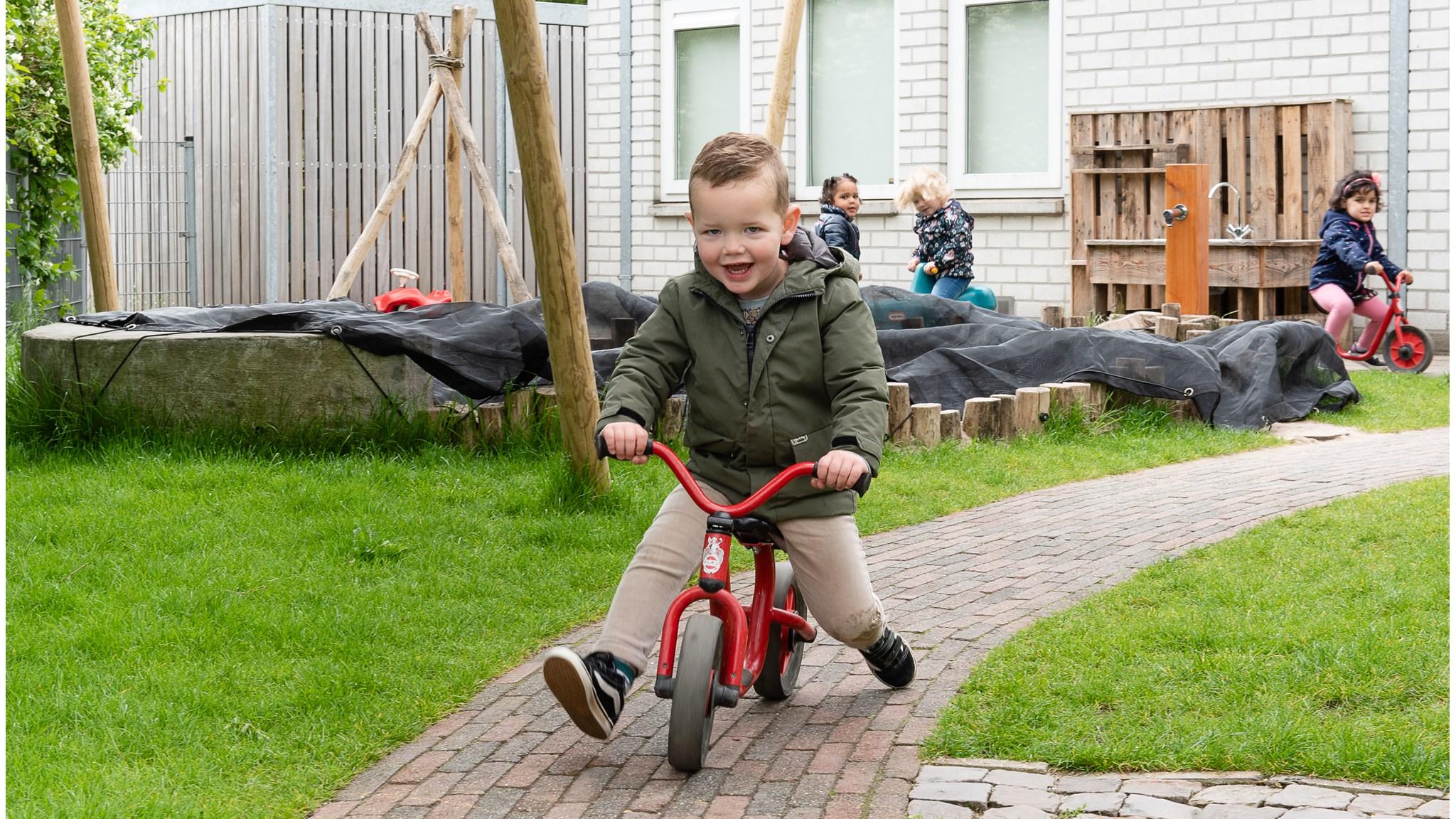 Een jongen rijdt blij op een rode loopfiets op het schoolplein van Integraal Kind Centrum Het Klokhuis in Duiven. Een jongen rijdt blij op een rode loopfiets op het schoolplein van Integraal Kind Centrum Het Klokhuis in Duiven.