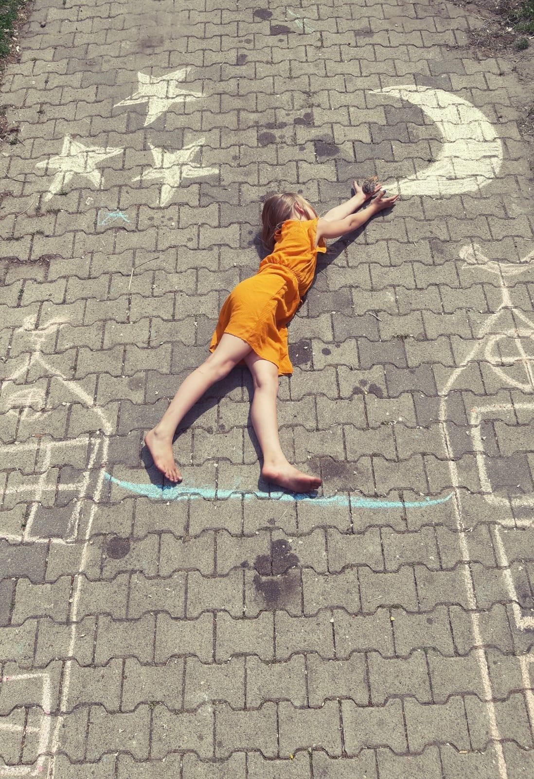 Een kind in een oranje jurk ligt op het schoolplein van KC 't Echtenest in Echten (Drenthe), omringd door met krijt getekende sterren en een maan. Een kind in een oranje jurk ligt op het schoolplein van KC 't Echtenest in Echten (Drenthe), omringd door met krijt getekende sterren en een maan.