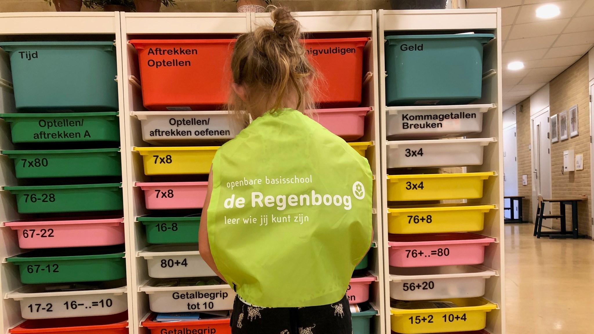 Een kind met een groene schort staat voor kleurrijke opbergdozen met leeractiviteiten in Openbare Basisschool de Regenboog in Voorthuizen. Een kind met een groene schort staat voor kleurrijke opbergdozen met leeractiviteiten in Openbare Basisschool de Regenboog in Voorthuizen.