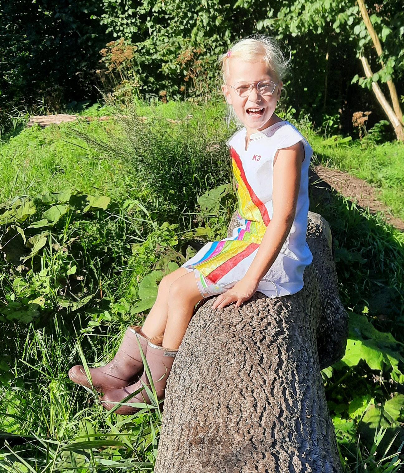 Een lachend meisje in vrolijke kleren zit op een boomstam in de natuur bij de St. Jozefschool in Achterveld (Utrecht). Een lachend meisje in vrolijke kleren zit op een boomstam in de natuur bij de St. Jozefschool in Achterveld (Utrecht).