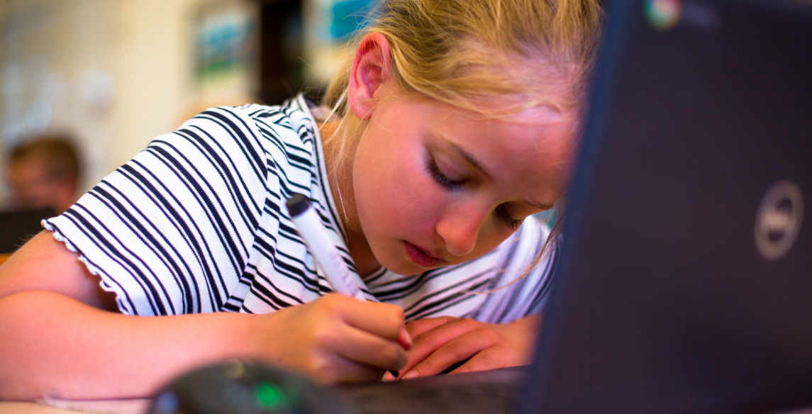 Een leerling van Basisschool Sint Jozef in Schipluiden is geconcentreerd aan het werk met haar laptop en schrijft iets op papier. Een leerling van Basisschool Sint Jozef in Schipluiden is geconcentreerd aan het werk met haar laptop en schrijft iets op papier.