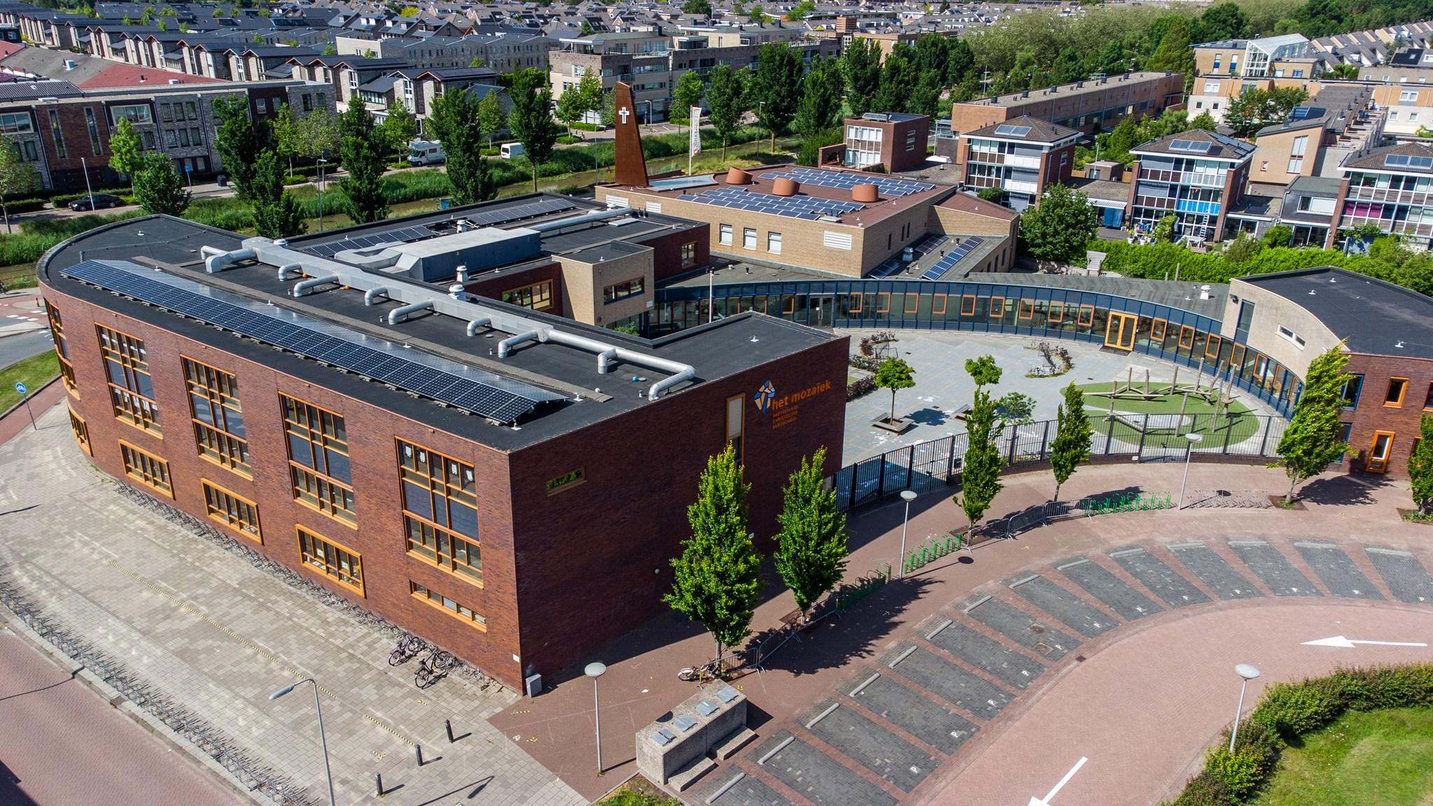 Een luchtfoto van basisschool Het Mozaïek in Alphen aan den Rijn met een modern schoolplein en duurzame zonnepanelen. Een luchtfoto van basisschool Het Mozaïek in Alphen aan den Rijn met een modern schoolplein en duurzame zonnepanelen.