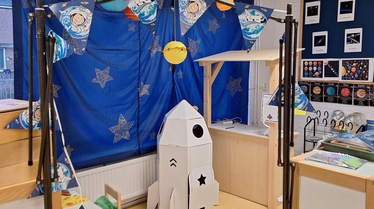 Een speelhoek in Openbare Basisschool De Piramide in Andijk, versierd met een ruimte-thema, inclusief een kartonnen raket en kleurrijke vlaggetjes. Een speelhoek in Openbare Basisschool De Piramide in Andijk, versierd met een ruimte-thema, inclusief een kartonnen raket en kleurrijke vlaggetjes.