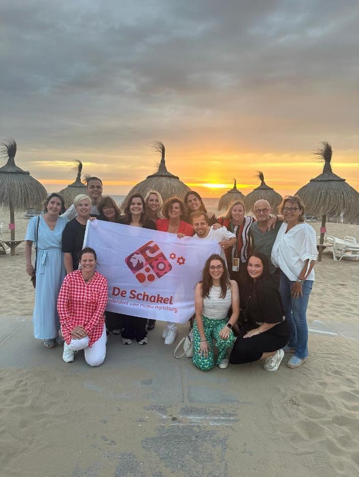 Foto van het team van P.C. Basisschool De Schakel in Rijnsburg met hun vlag op het strand tijdens zonsondergang. Foto van het team van P.C. Basisschool De Schakel in Rijnsburg met hun vlag op het strand tijdens zonsondergang.