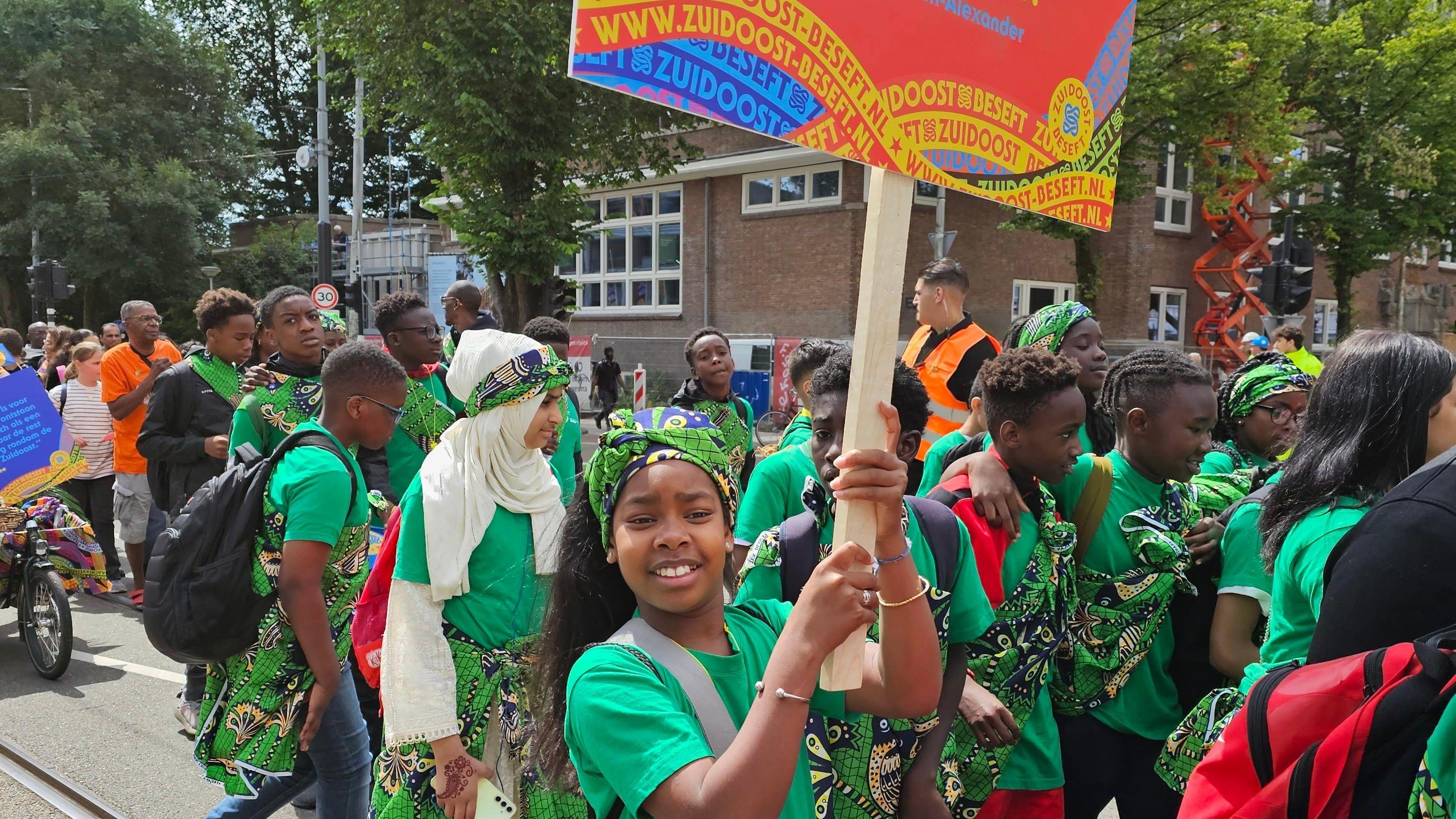 Groep gelukkige kinderen van IKC Knotwilg in Amsterdam, gekleed in groene shirts en kleurrijke doeken, tijdens een parade. Groep gelukkige kinderen van IKC Knotwilg in Amsterdam, gekleed in groene shirts en kleurrijke doeken, tijdens een parade.