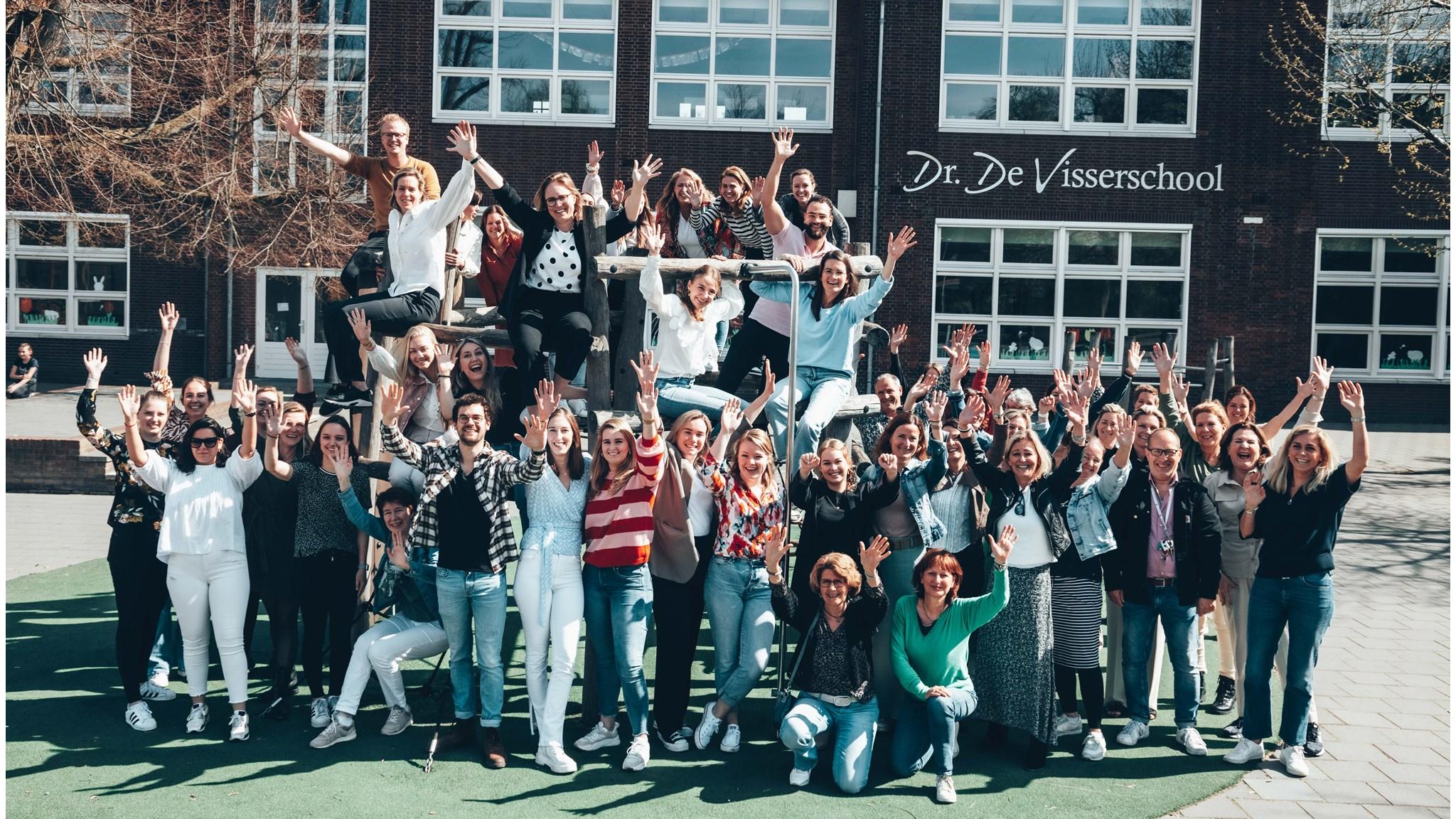 Groepsfoto van het personeel van Dr. De Visserschool in Breda, poserende op het schoolplein en met opgestoken handen.