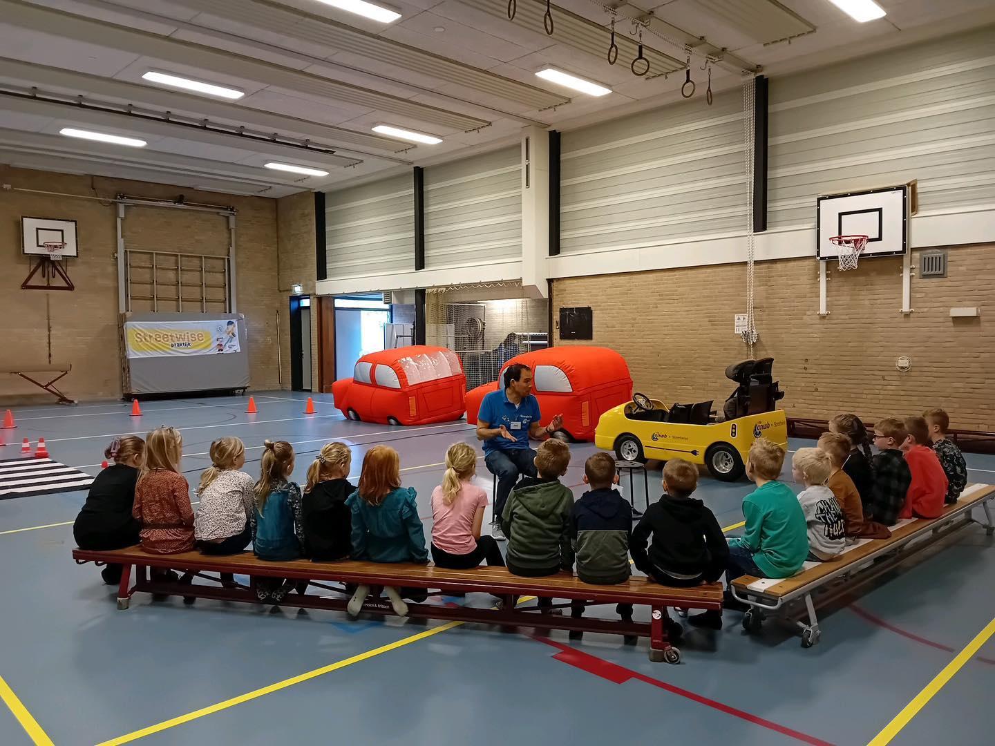 In de gymzaal van It Kruirêd in Mûnein zitten kinderen te luisteren naar een instructeur over verkeersveiligheid met speelauto's en verkeershindernissen.