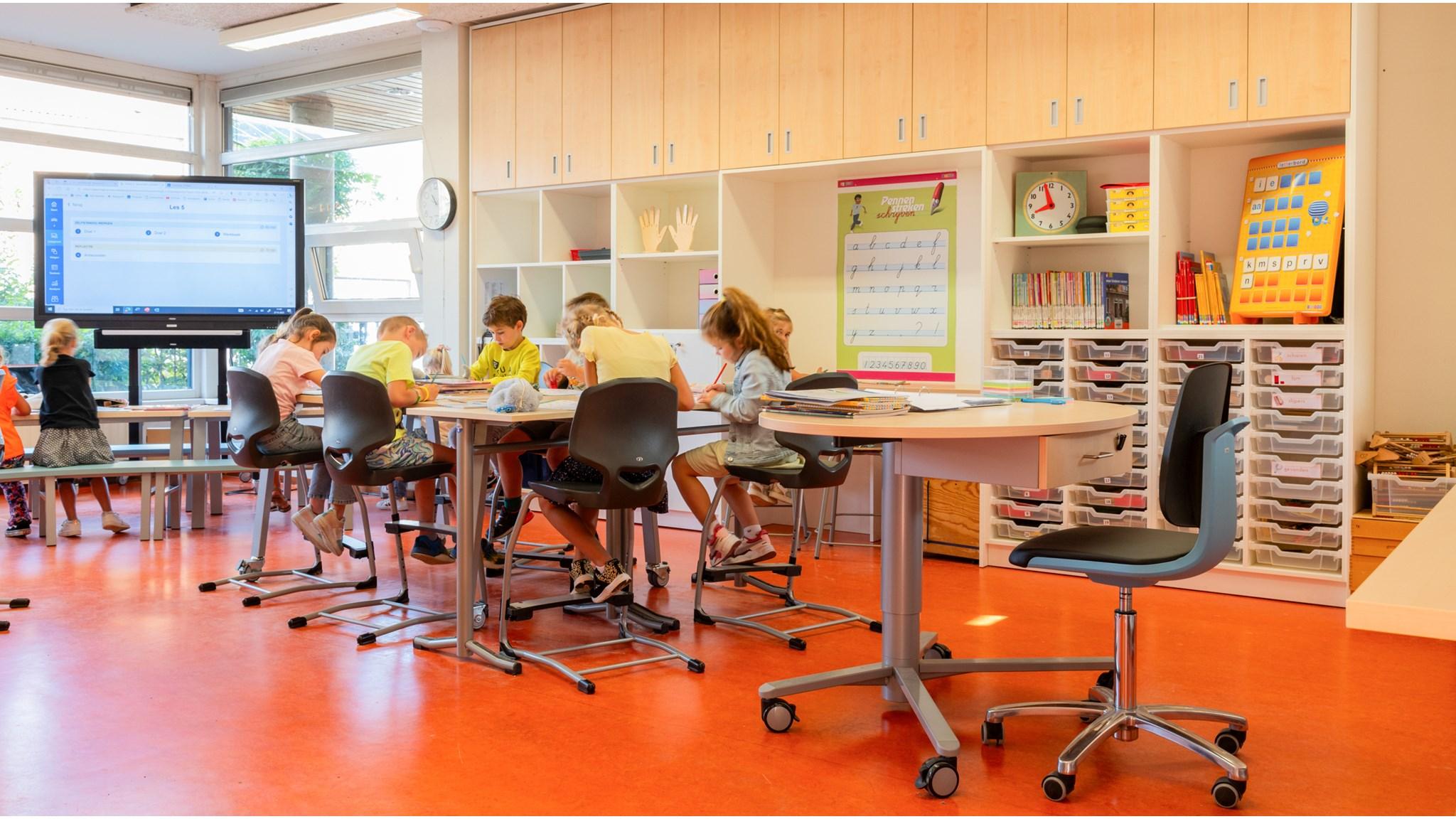 In het Kindcentrum Amby in Maastricht zitten kinderen geconcentreerd aan hun werktafels in een kleurrijke klaslokaal. In het Kindcentrum Amby in Maastricht zitten kinderen geconcentreerd aan hun werktafels in een kleurrijke klaslokaal.