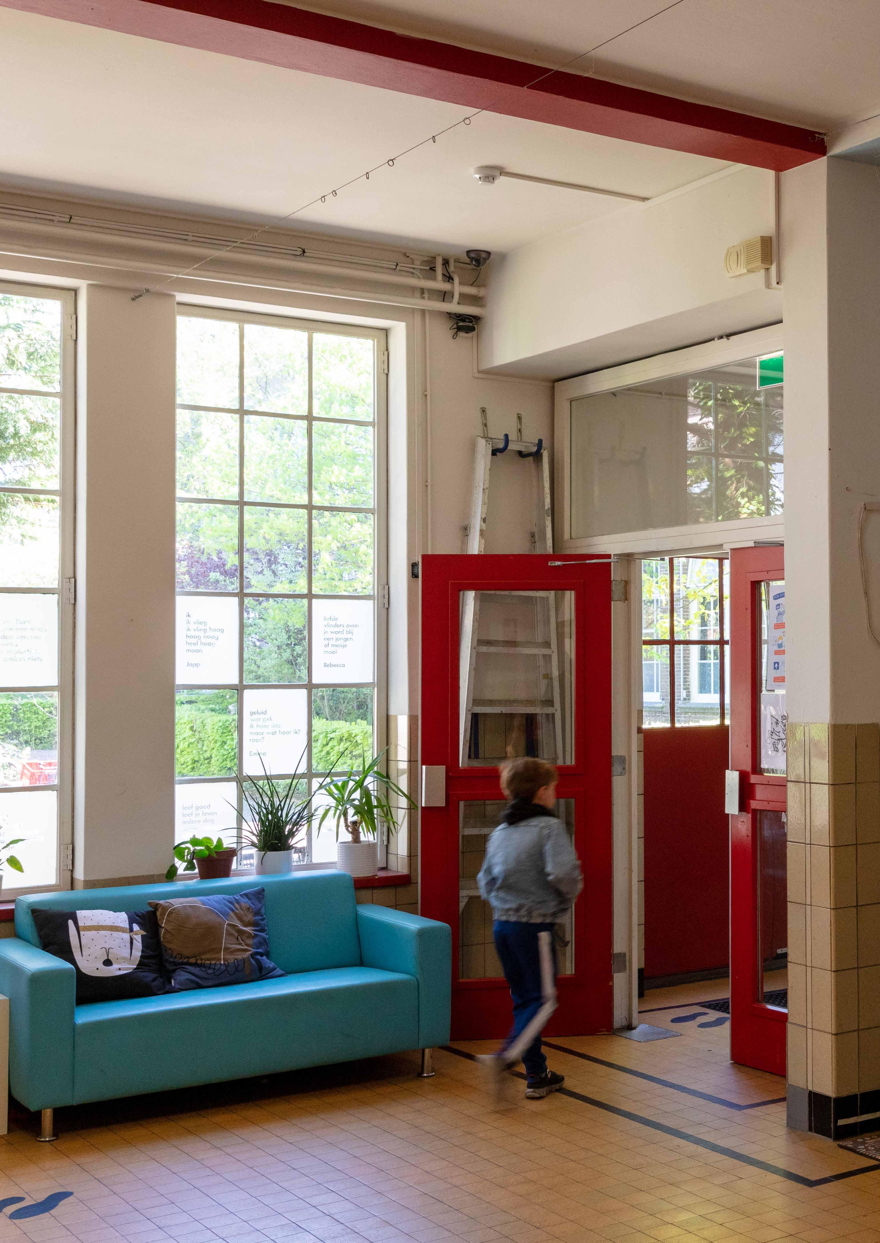 Interieur van de Openbare Basisschool Bornwaterschool in Bloemendaal met een kind dat voorbijloopt bij een kleurrijke bank en grote ramen. Interieur van de Openbare Basisschool Bornwaterschool in Bloemendaal met een kind dat voorbijloopt bij een kleurrijke bank en grote ramen.