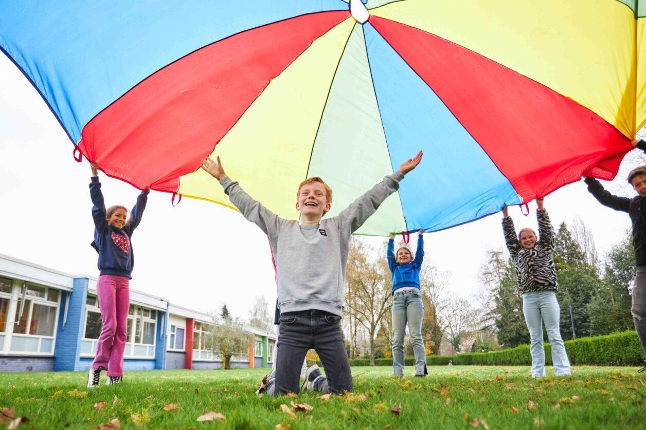 Kinderen spelen samen op het schoolplein van Basisschool De Bron in Sprang-Capelle met een grote parachute. Kinderen spelen samen op het schoolplein van Basisschool De Bron in Sprang-Capelle met een grote parachute.