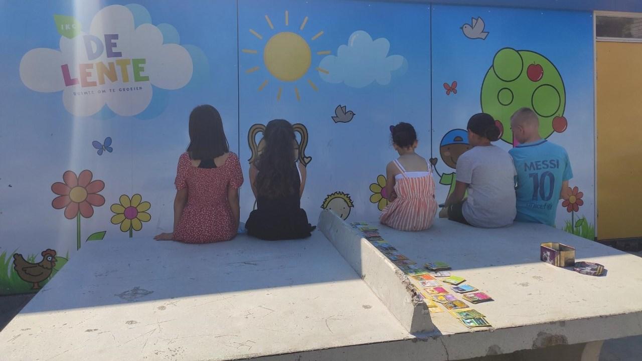 Kinderen zitten op het schoolplein van IKC De Lente in Abbenes, terwijl ze naar de kleurrijke muurschildering kijken. Kinderen zitten op het schoolplein van IKC De Lente in Abbenes, terwijl ze naar de kleurrijke muurschildering kijken.