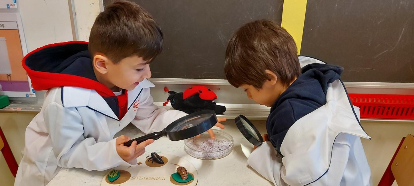 Twee kinderen in laboratoriumjassen onderzoeken met vergrootglazen een schaaltje in klaslokaal van De Kleine Wereld in Den Haag. Twee kinderen in laboratoriumjassen onderzoeken met vergrootglazen een schaaltje in klaslokaal van De Kleine Wereld in Den Haag.