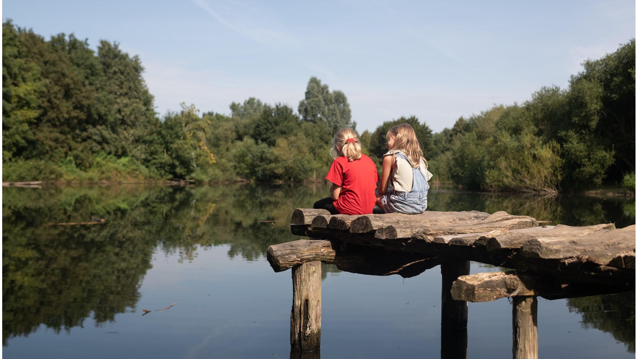 Twee kinderen op een houten pier bij IKC De Kleine Reus in Amsterdam, zittend aan een spiegelend water. Twee kinderen op een houten pier bij IKC De Kleine Reus in Amsterdam, zittend aan een spiegelend water.