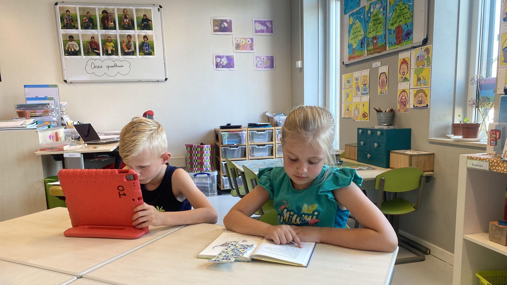 Twee kinderen zitten aan een tafel in de klas van obs Prinses Wilhelmina in Lexmond, één leest een boek en de ander kijkt op een tablet.
