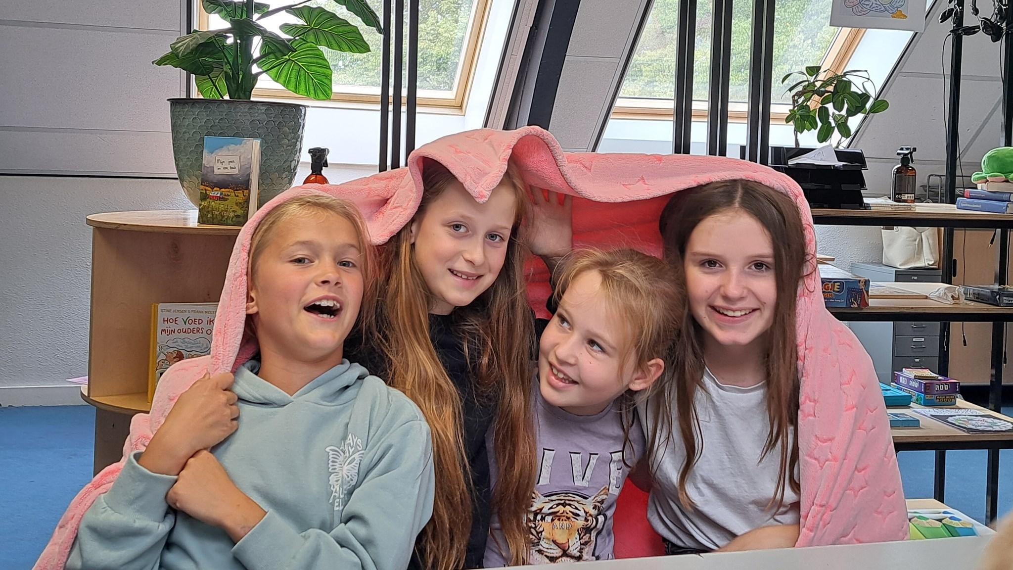 Vier blije kinderen zitten samen onder een roze deken op Basisschool De Heihorst in Heibloem. Vier blije kinderen zitten samen onder een roze deken op Basisschool De Heihorst in Heibloem.